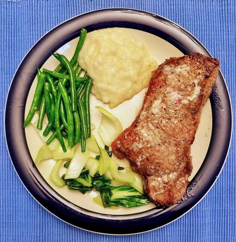 A plate of New York strip steak with butter, mashed potatoes, green beans and bok choy